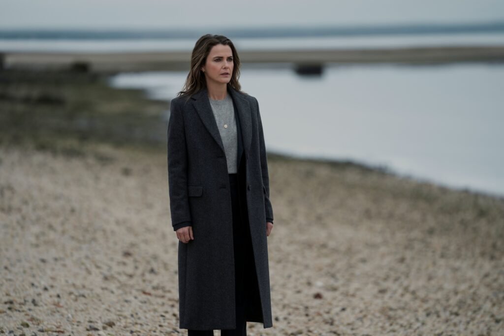 "Woman in a long dark coat standing alone on a rocky beach, with calm water and an overcast sky in the background."