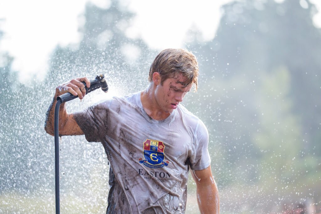 "A young man outdoors sprays himself with water from a hose to clean off mud, wearing a dirty t-shirt with a crest in a natural, sunlit environment, suggesting he has just finished a muddy outdoor activity or sport."