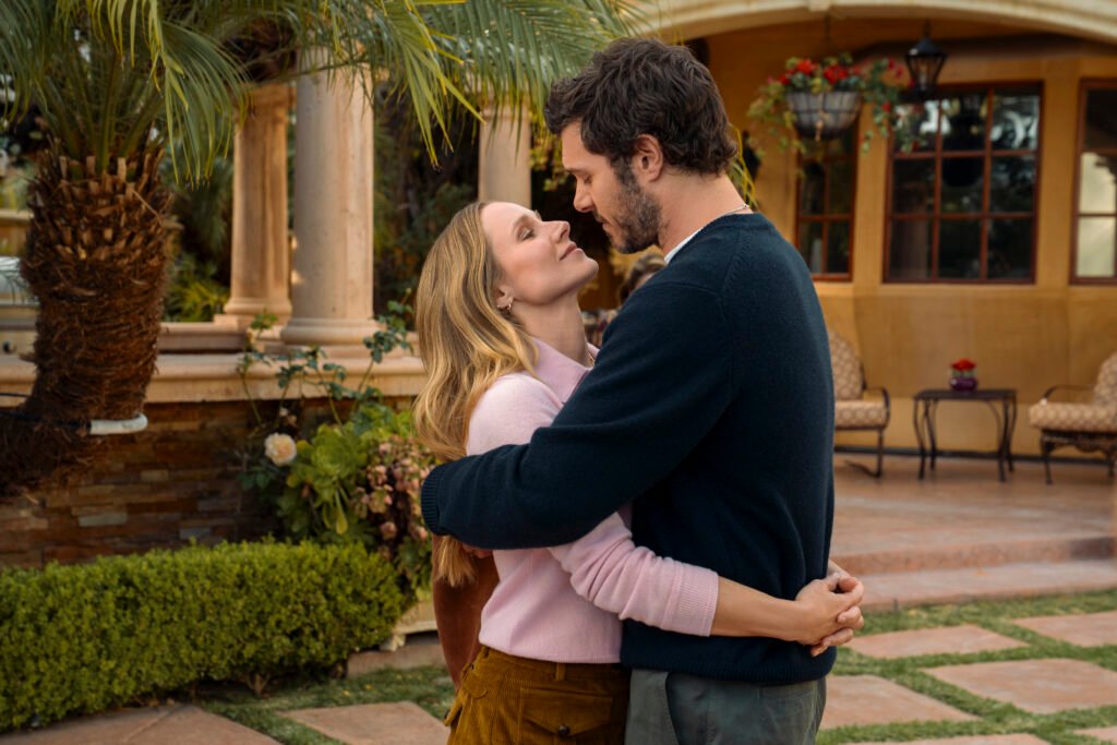 "A man and woman embrace and look into each other's eyes in a lush garden setting with palm trees, manicured bushes, and an elegant patio with chairs and columns in the background."